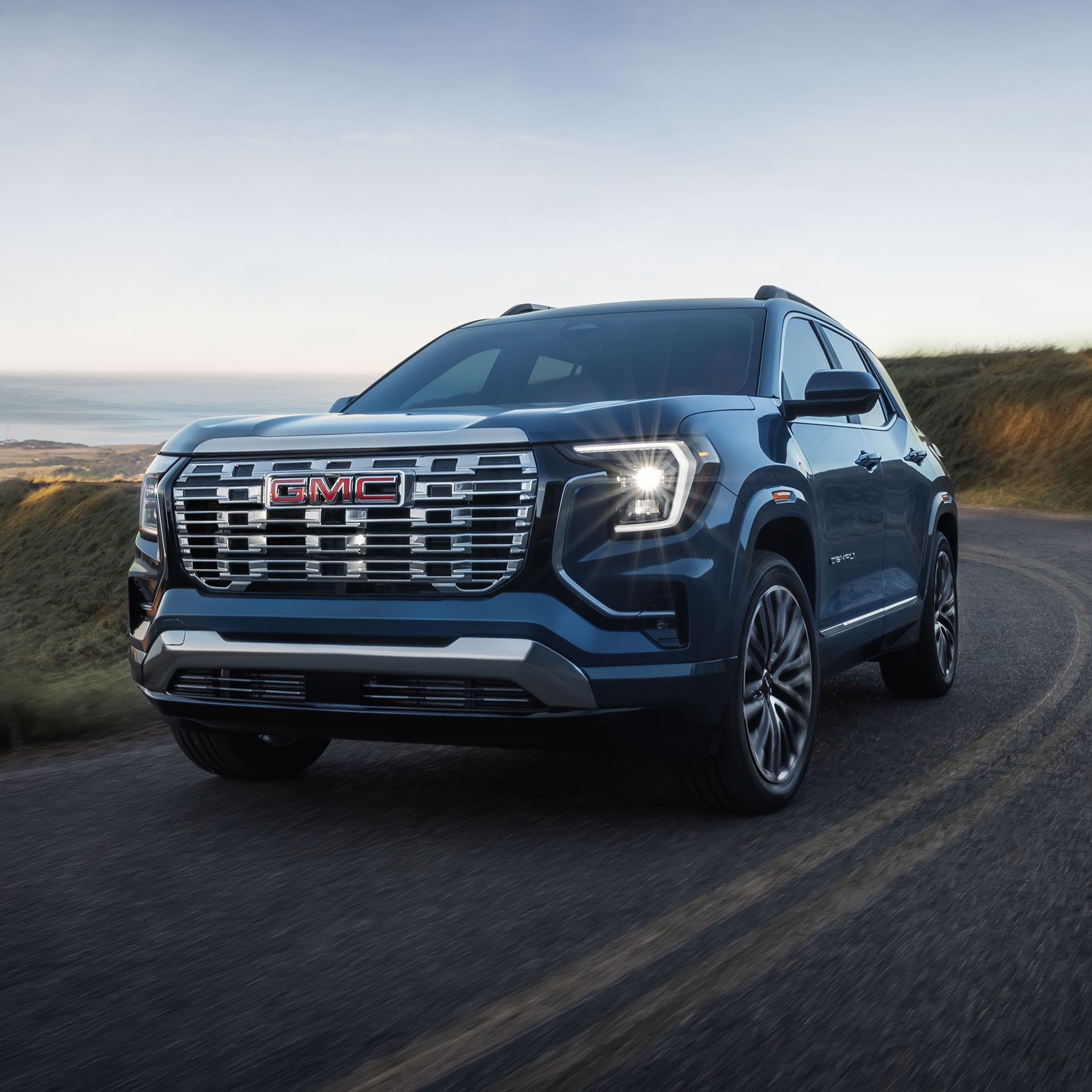 Un SUV GMC Terrain Denali bleu foncé 2027 roulant sur une route sinueuse surplombant un paysage côtier pittoresque.