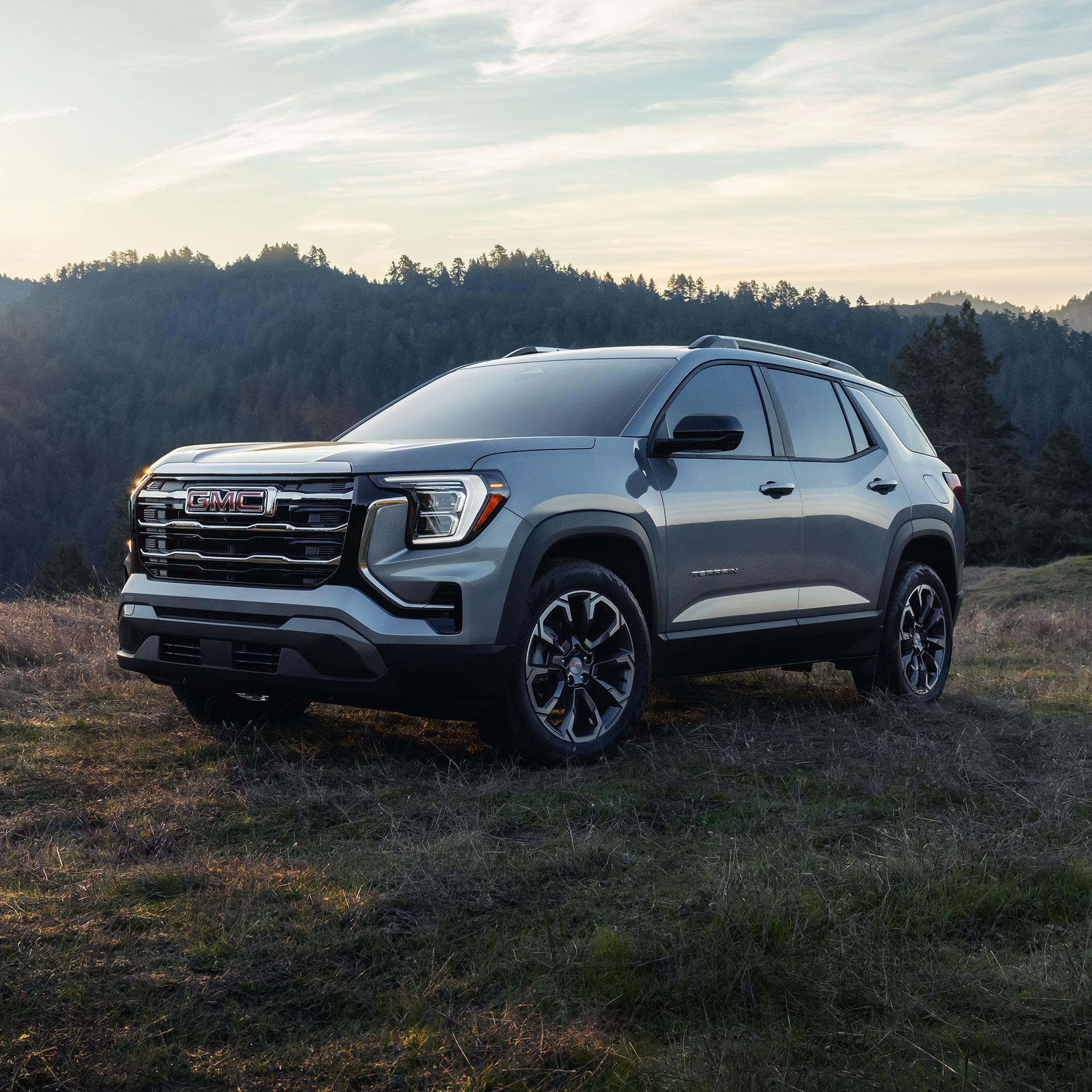 Un SUV GMC Terrain gris clair 2027 garé sur une colline herbeuse avec la lumière du soleil brillant sur les montagnes boisées.