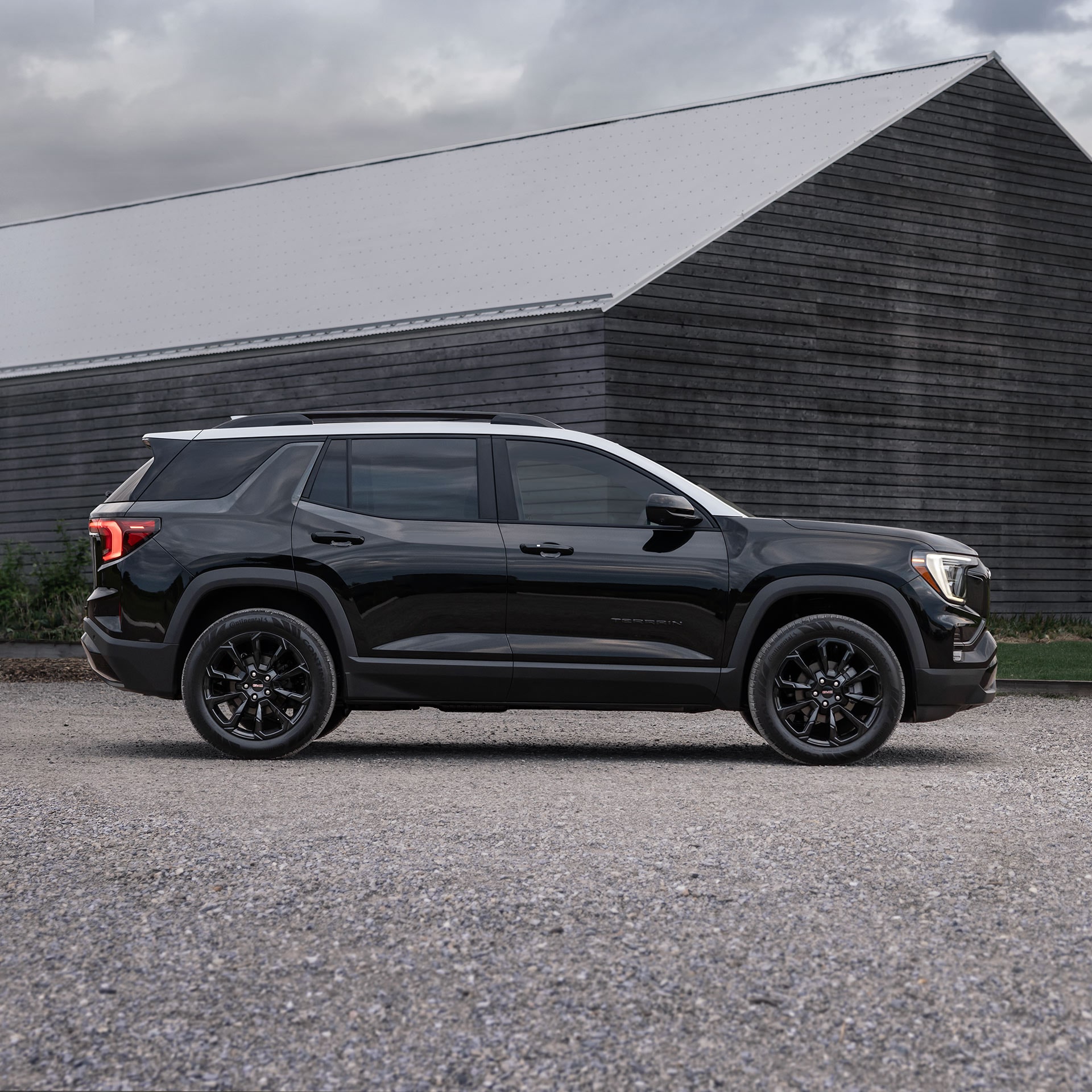 Un SUV GMC Terrain noir 2027 avec un toit blanc garé sur du gravier à côté d’un bâtiment moderne en bois.