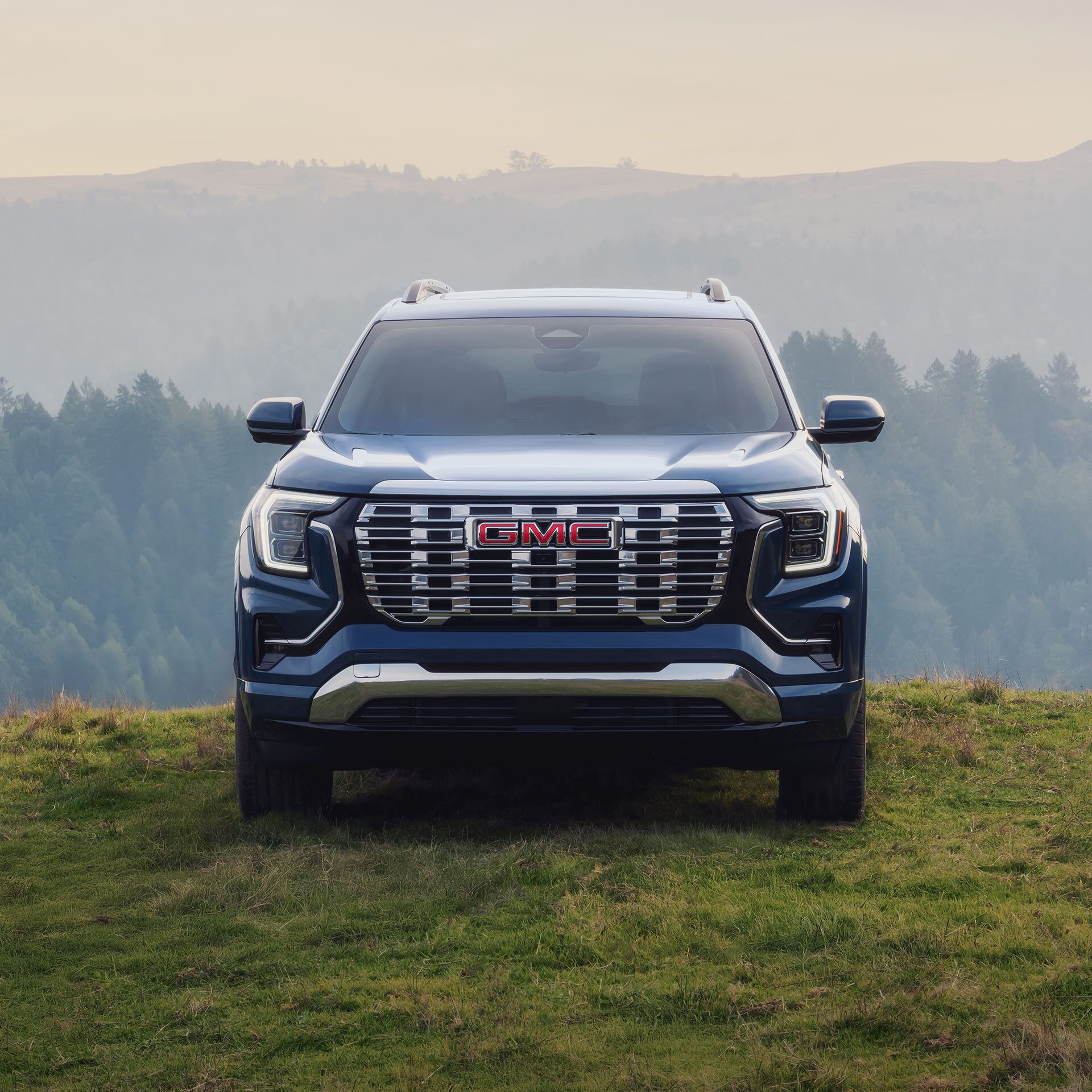 A Blue 2027 GMC Terrain SUV Parked on a Grassy Hill with Forested Mountains in the Background.