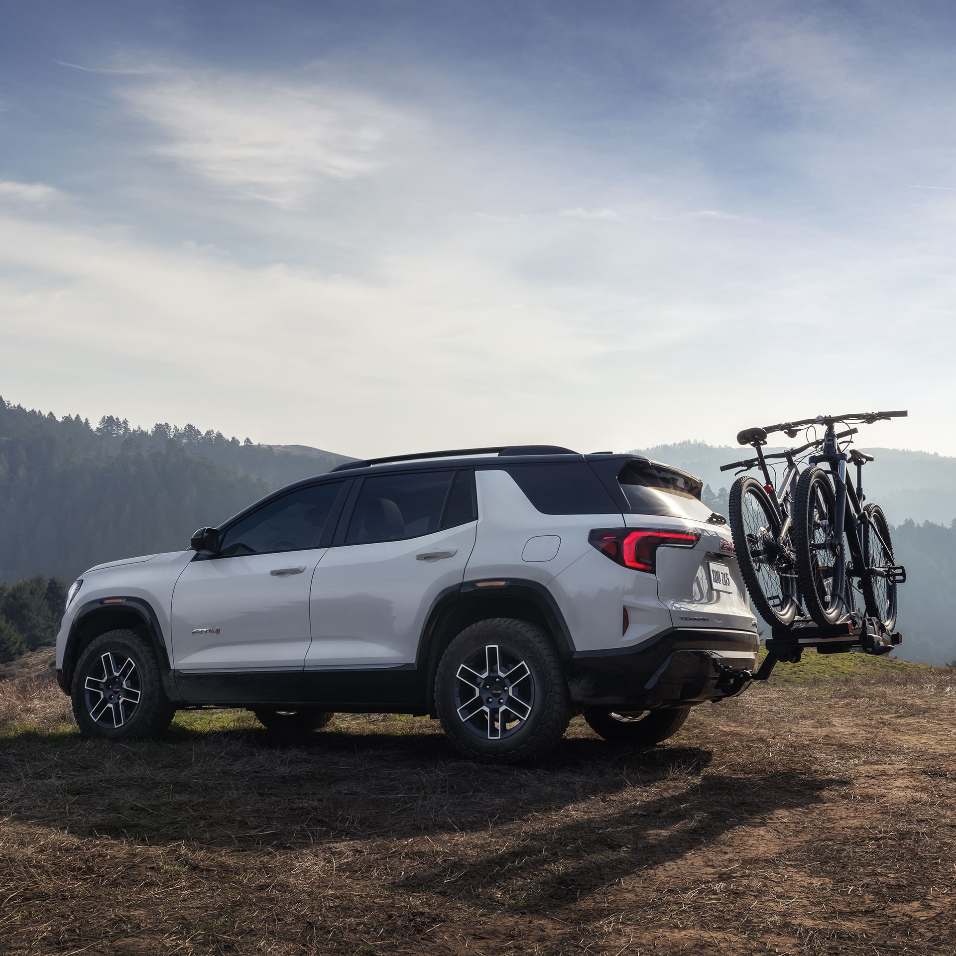 Un GMC Terrain Acadia AT4 blanc 2027 garé sur une colline herbeuse avec deux VTT posés sur un porte-vélos arrière.