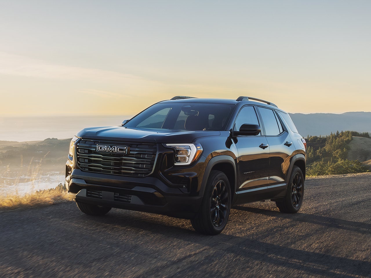 Une GMC Terrain Acadia AT4 noire 2027 garée sur une route sinueuse au coucher du soleil, avec un paysage brumeux en arrière-plan.