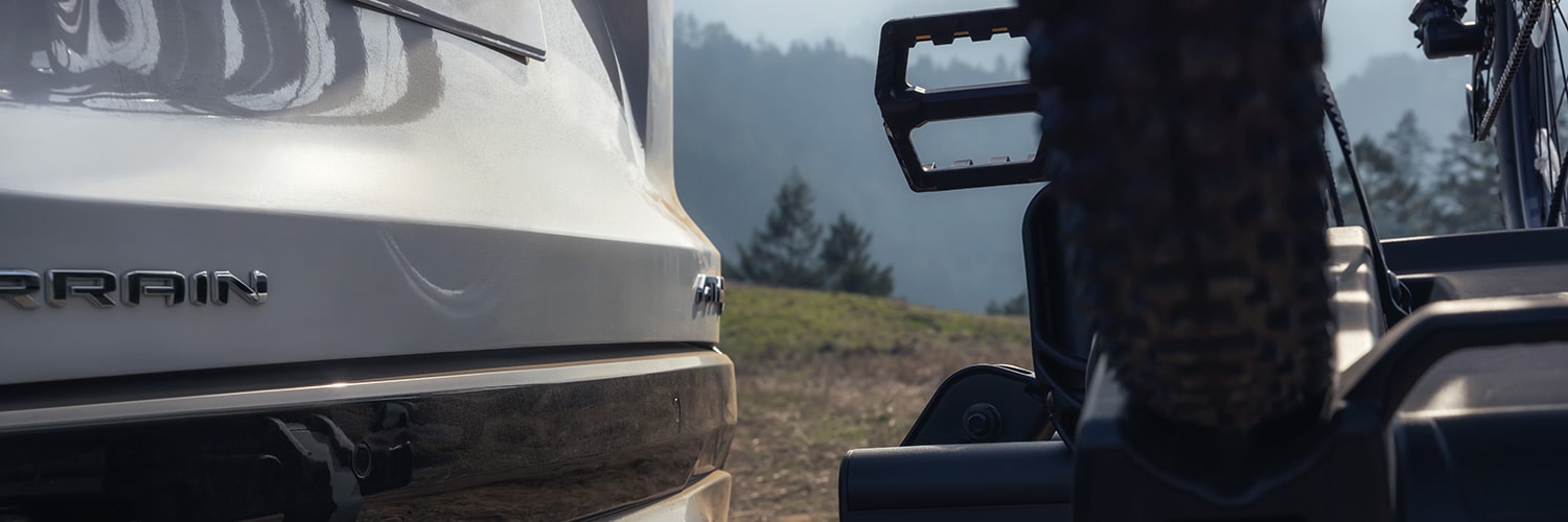 Vue rapprochée du pare-chocs arrière d’un SUV terrain GMC blanc 2027 avec un porte-vélos tenant une roue de vélo.