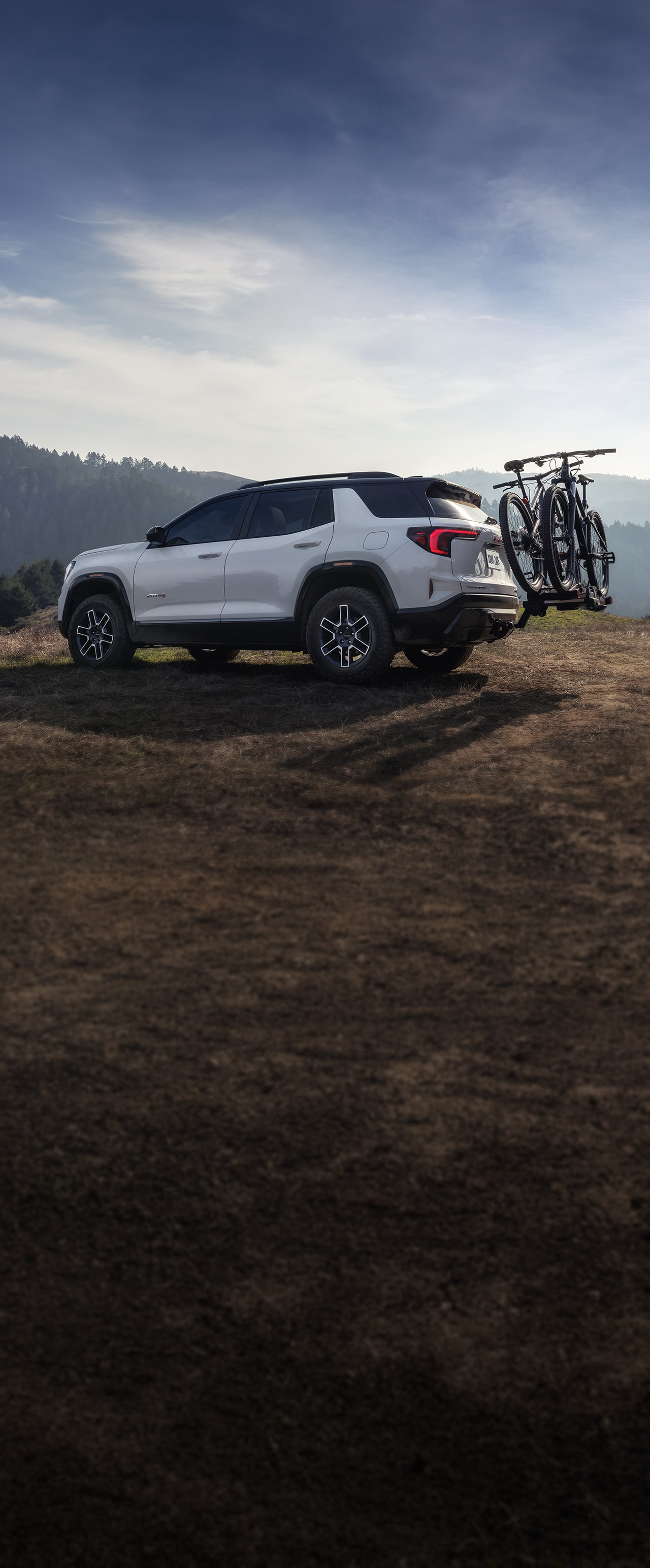 Un GMC Terrain Acadia AT4 blanc 2027 garé sur une colline herbeuse avec deux VTT posés sur un porte-vélos arrière.