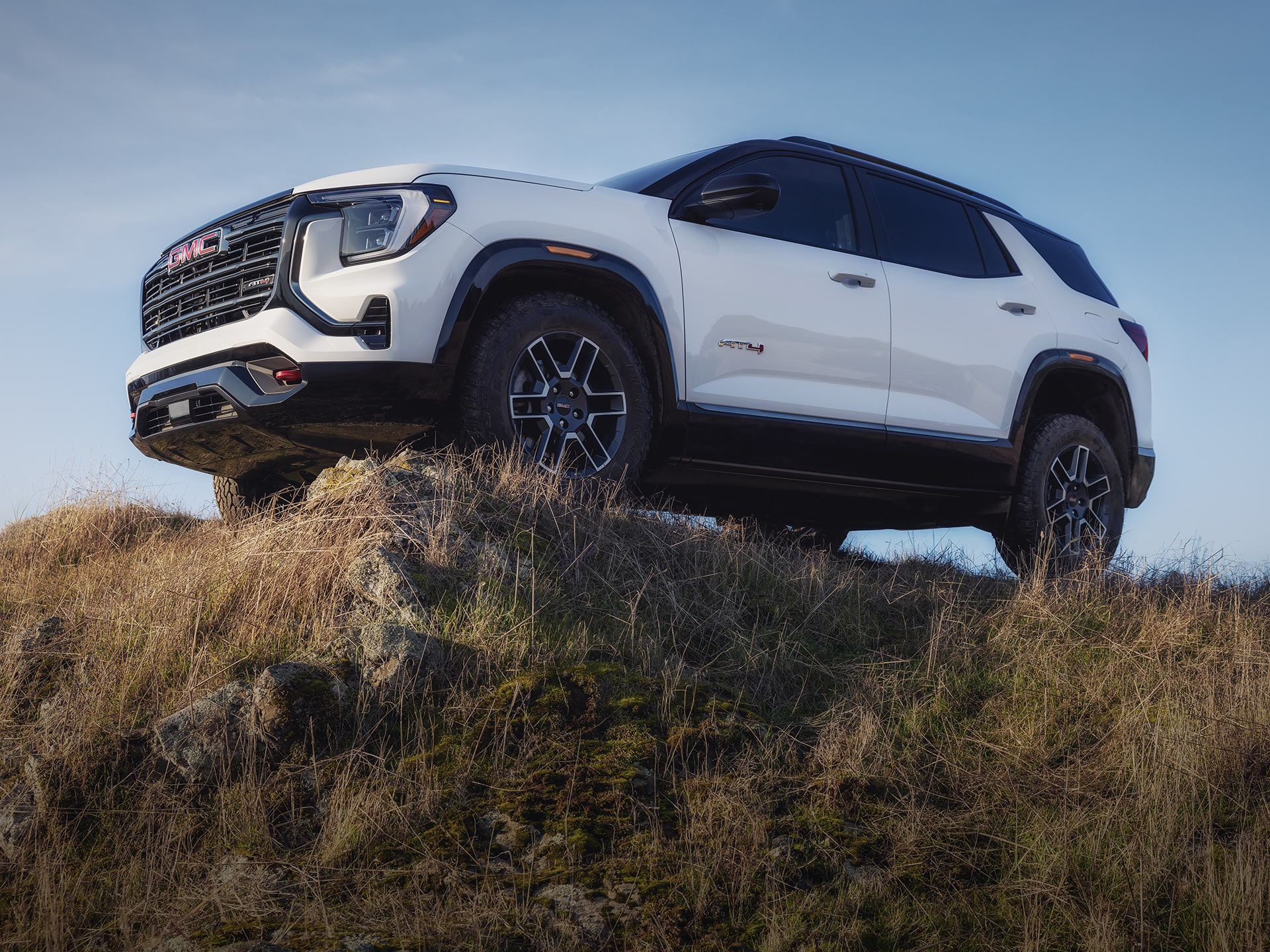 Un SUV AT4 GMC Terrain blanc 2027 perché sur une colline rocheuse et herbeuse sous un ciel bleu clair.