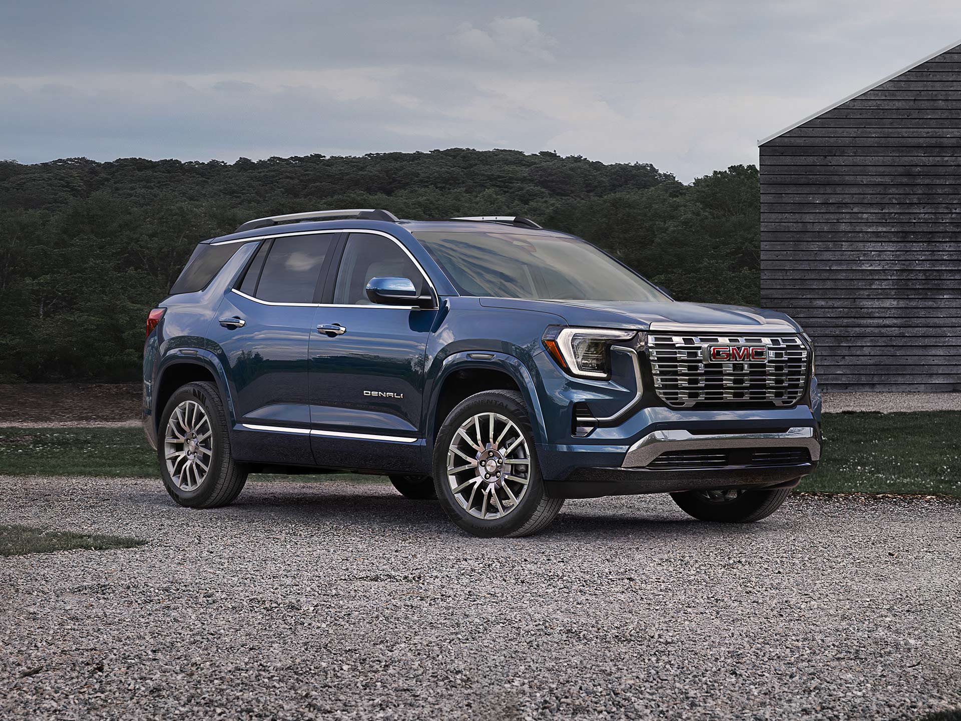 Un SUV GMC Terrain Denali bleu 2027 s’est garé sur un terrain de gravier devant un bâtiment en bois et des arbres sous un ciel nuageux.
