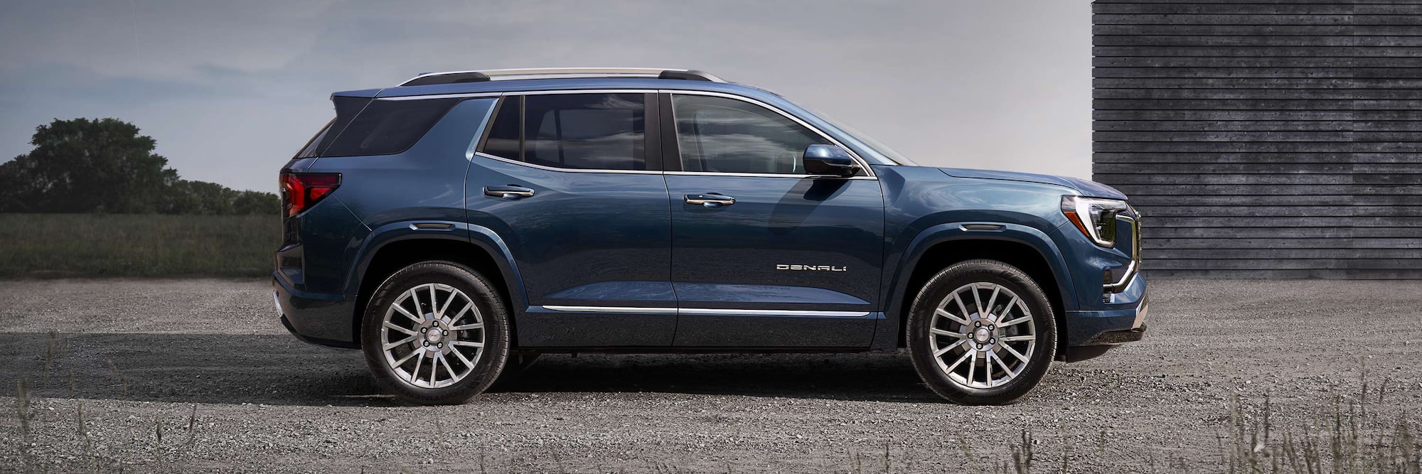 Vue de côté d’un SUV Denali GMC Terrain bleu 2027 garé sur une surface de gravier à côté d’une structure en bois.