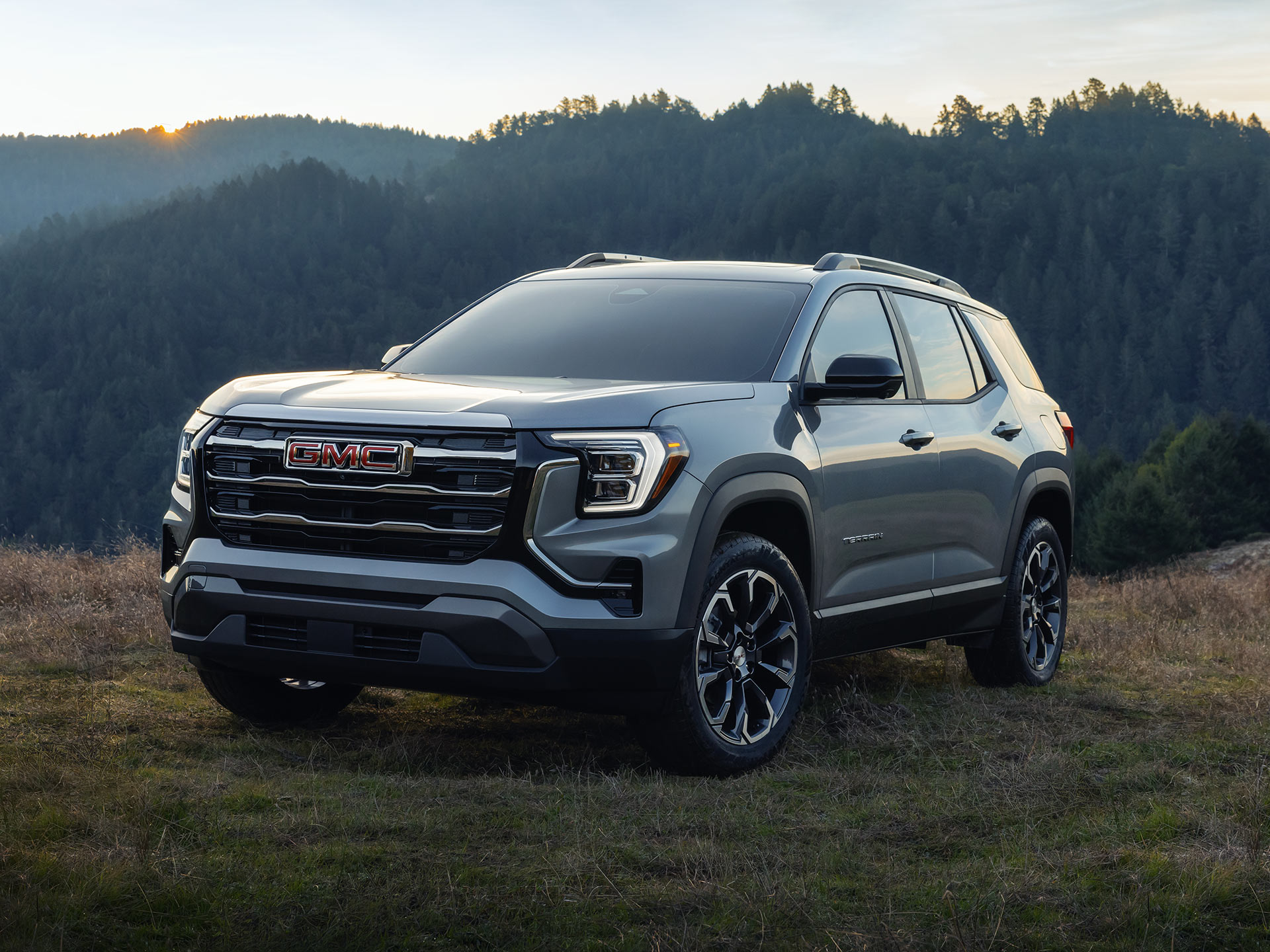 Un SUV Denali GMC Terrain 2027 gris garé dans un champ herbeux avec des collines boisées et un coucher de soleil en arrière-plan.
