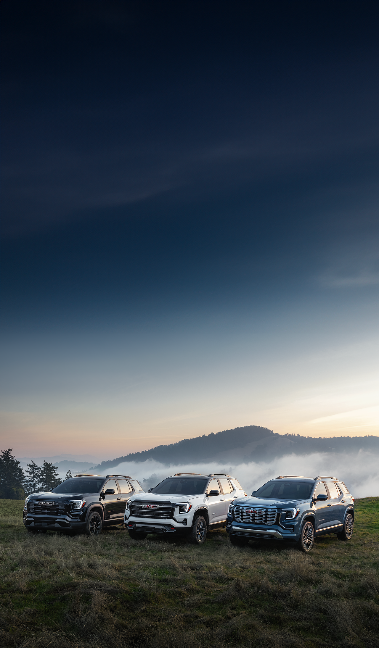 Trois SUV GMC Acadia, un noir, un blanc et un bleu, garés sur une colline herbeuse avec des montagnes brumeuses en arrière-plan.