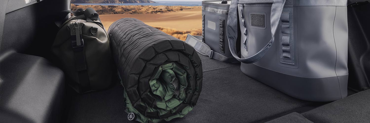 Équipement de camping incluant un matelas de couchage et un sac glacière dans le coffre d’un SUV GMC Terrain 2027 avec un paysage côtier au-delà.