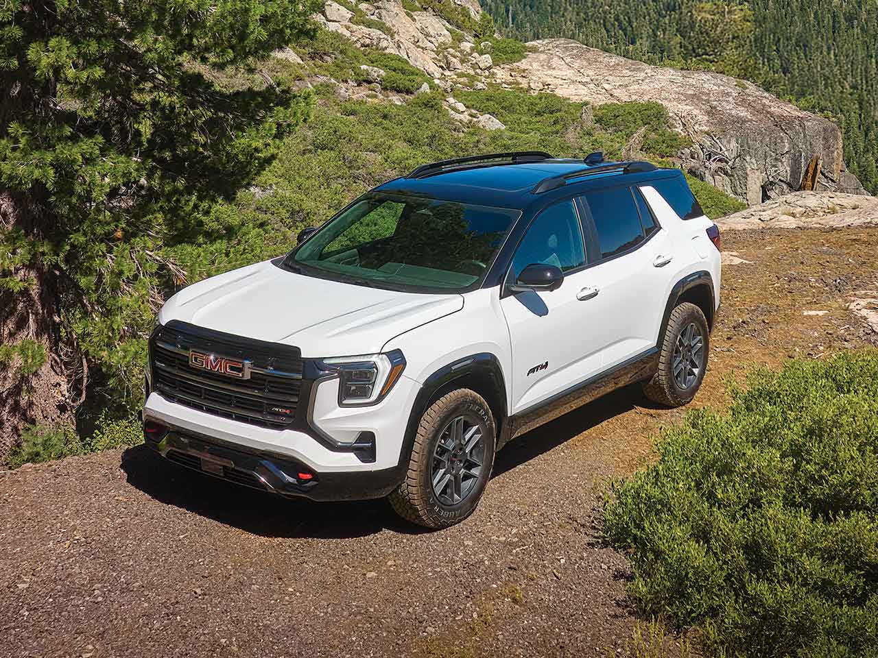 Un SUV AT4 GMC Terrain blanc 2027 garé sur un sentier de terre à côté d’arbres verts et de terrain rocailleux sous un ciel bleu clair.
