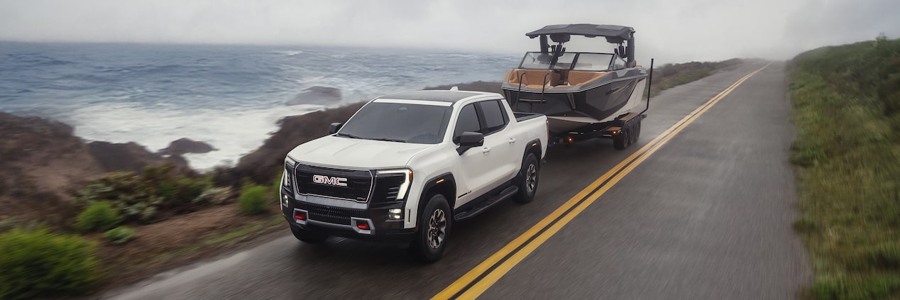 Vue latérale avant du pick-up GMC Sierra EV 2026 qui remorque un bateau sur une route au bord de la mer.