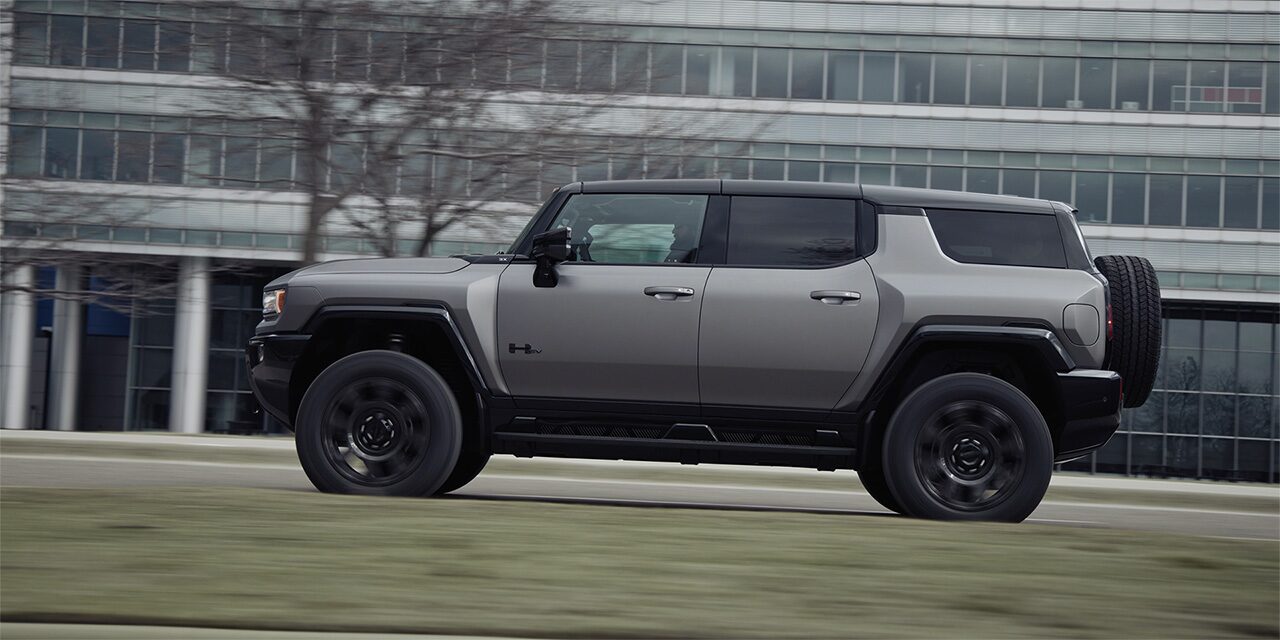 Vue latérale avant du VUS GMC Hummer EV 2026 en mouvement à côté d’un bâtiment.