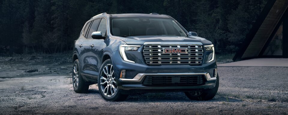 GMC Acadia Denali 2024 stationné seul sur du gravier devant des arbres.