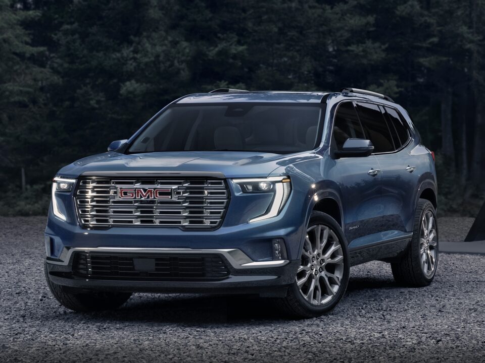 Vue latérale avant du GMC Acadia Denali 2024 stationné sur du gravier devant des arbres.