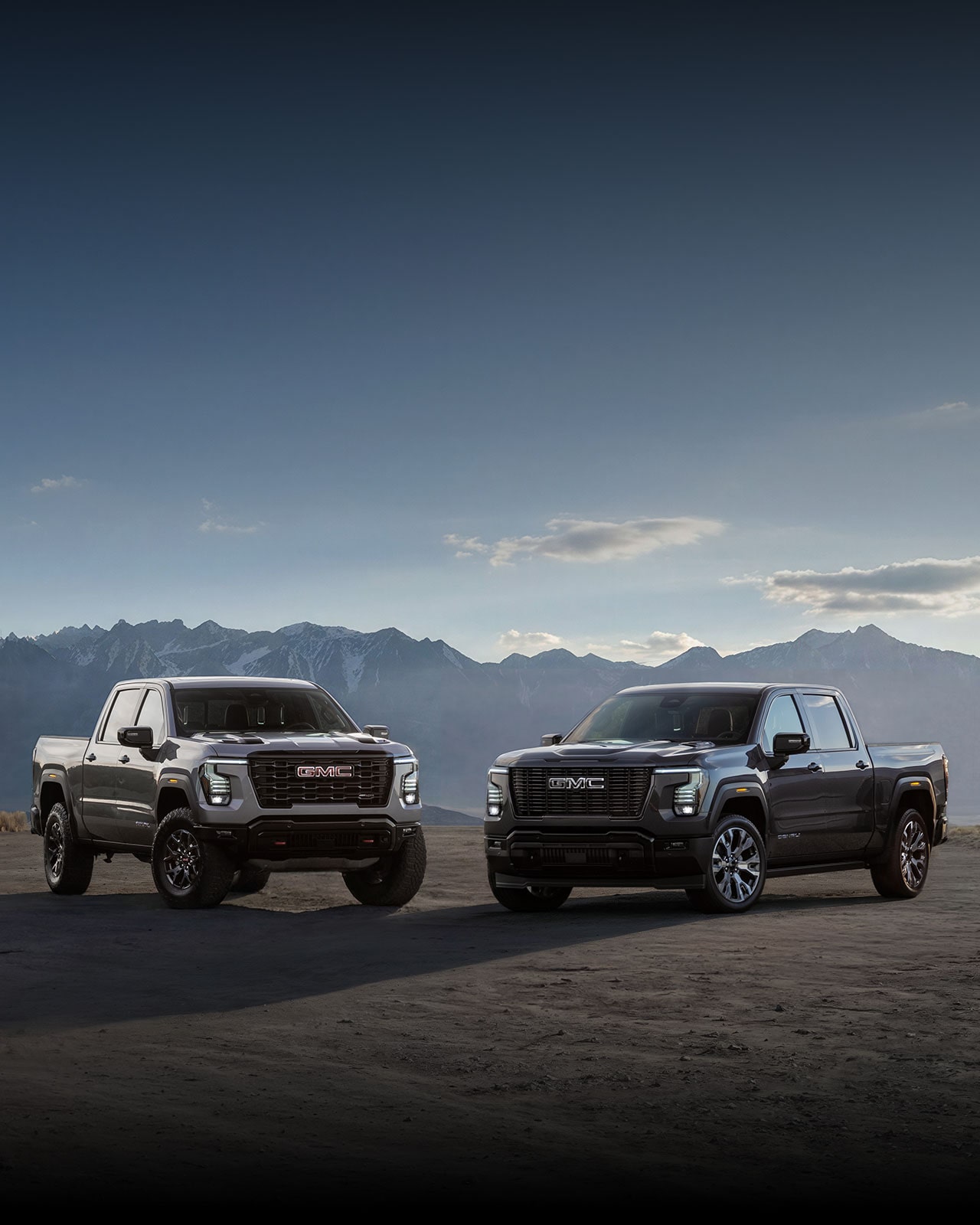 Two next-generation 2027 GMC Sierra 1500 pickup trucks parked side by side on an open road with a mountainous landscape in the background.