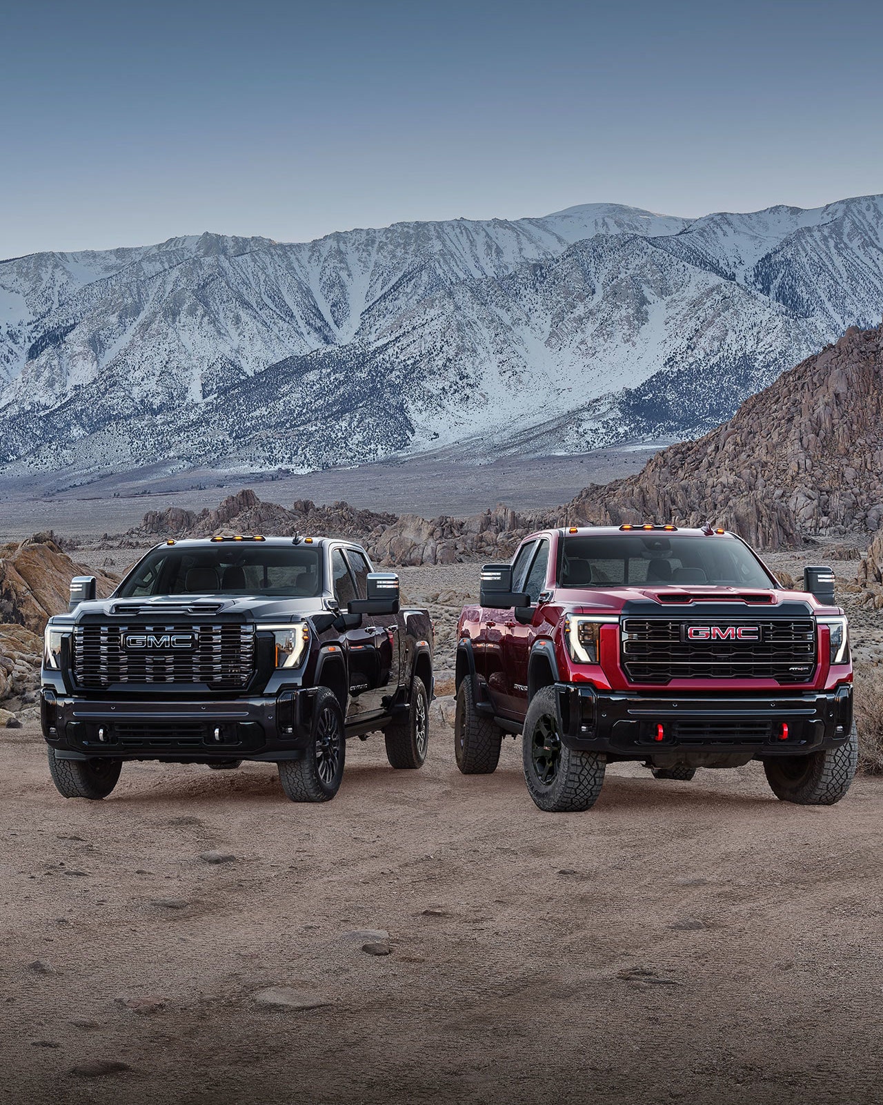 Two 2027 GMC Sierra HD Trucks, One Black and One Red, with Off-Road Tires and Red Recovery Hooks Parked Side By Side On a Rocky Terrain with Snowy Mountains in the Background