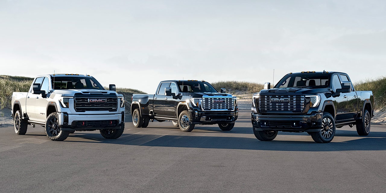 GMC Sierra HD AT4 blanc, Sierra HD Denali gris foncé et Sierra HD Denali Ultimate noir roulant sur une route bordée de dunes de sable.