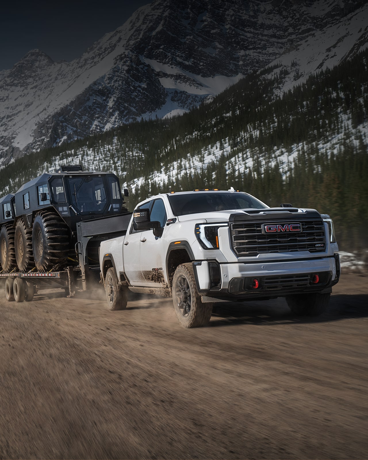 GMC Sierra HD AT4X blanc remorquant une remorque hors route dans un désert rocheux avec des montagnes à l’arrière-plan.