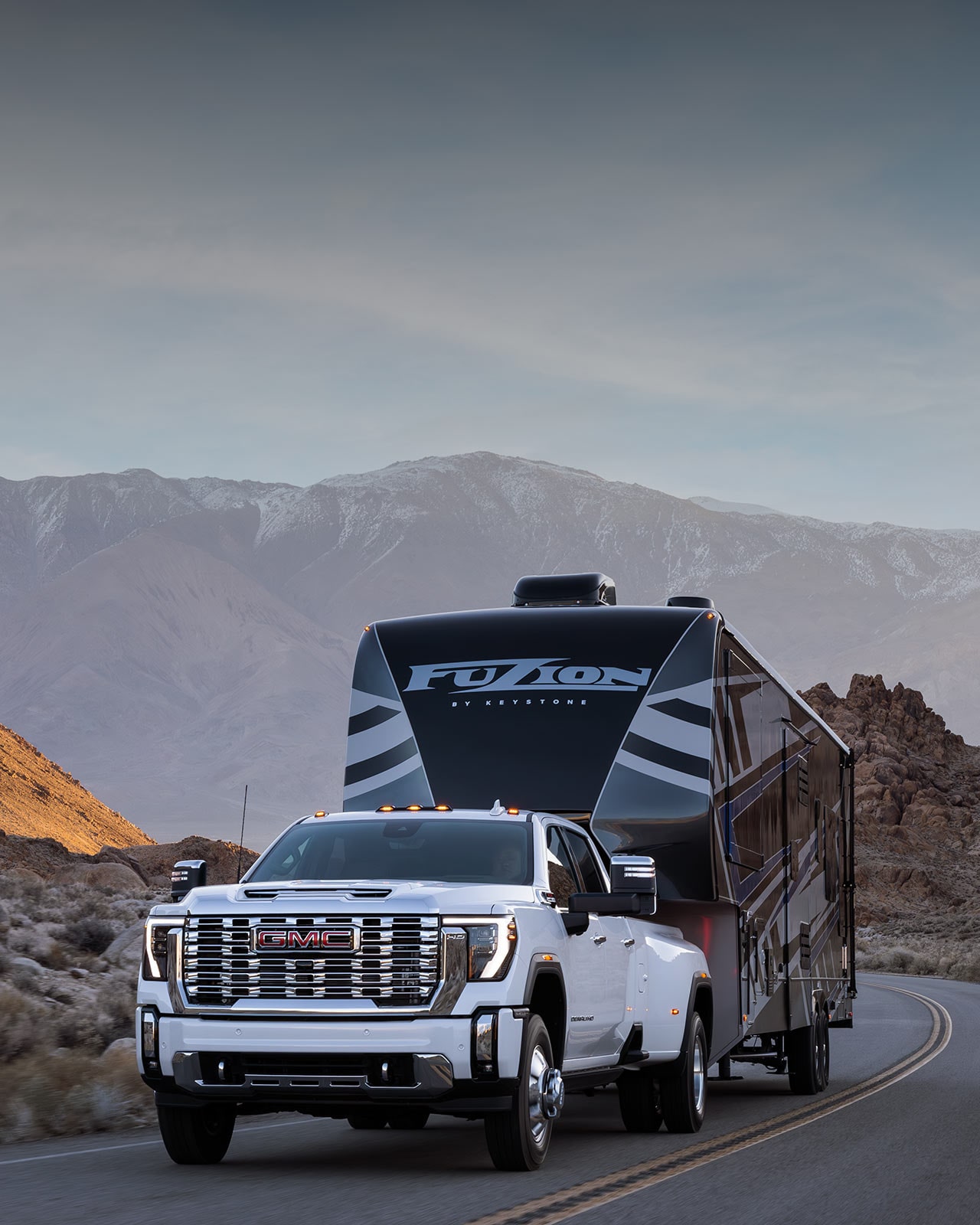 GMC Sierra HD 2027 blanc à roues arrière jumelées remorquant une grande roulotte à sellette noire et blanche dans un décor désertique montagneux.