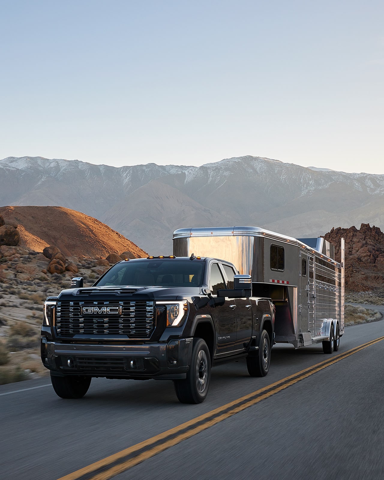 GMC Sierra HD 2027 noir remorquant une remorque à chevaux argentée sur une route asphaltée dans un paysage rocheux et montagneux.