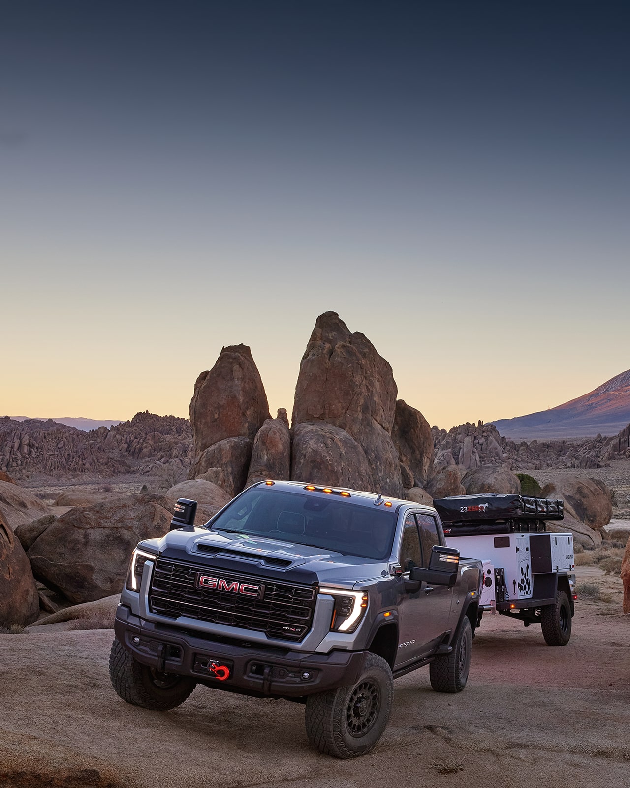 GMC Sierra HD 2027 gris foncé avec crochets de remorquage rouges et pneus tout-terrain, remorquant une petite remorque d’aventure blanche sur un terrain rocheux.