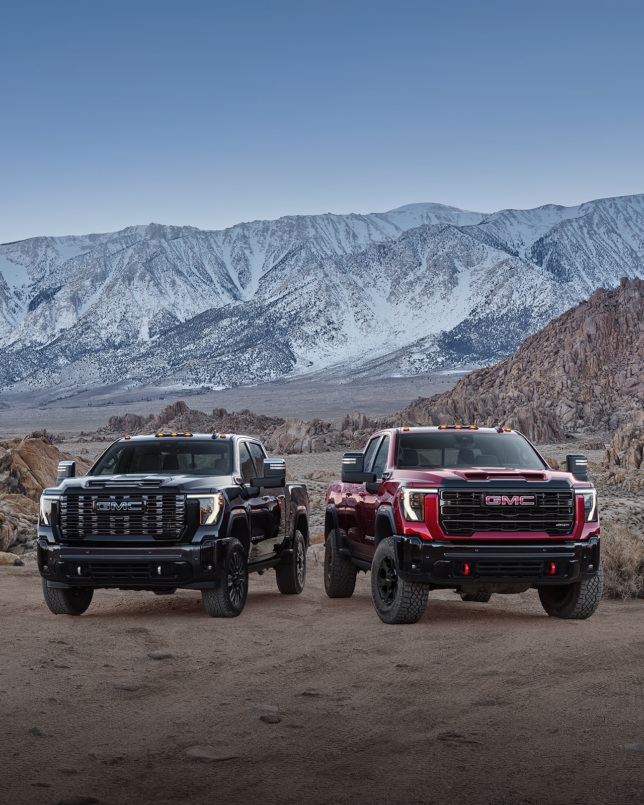 Camions GMC Sierra HD 2027 noir et rouge avec pneus tout-terrain et crochets de remorquage rouges sur un terrain rocheux, devant des montagnes enneigées.