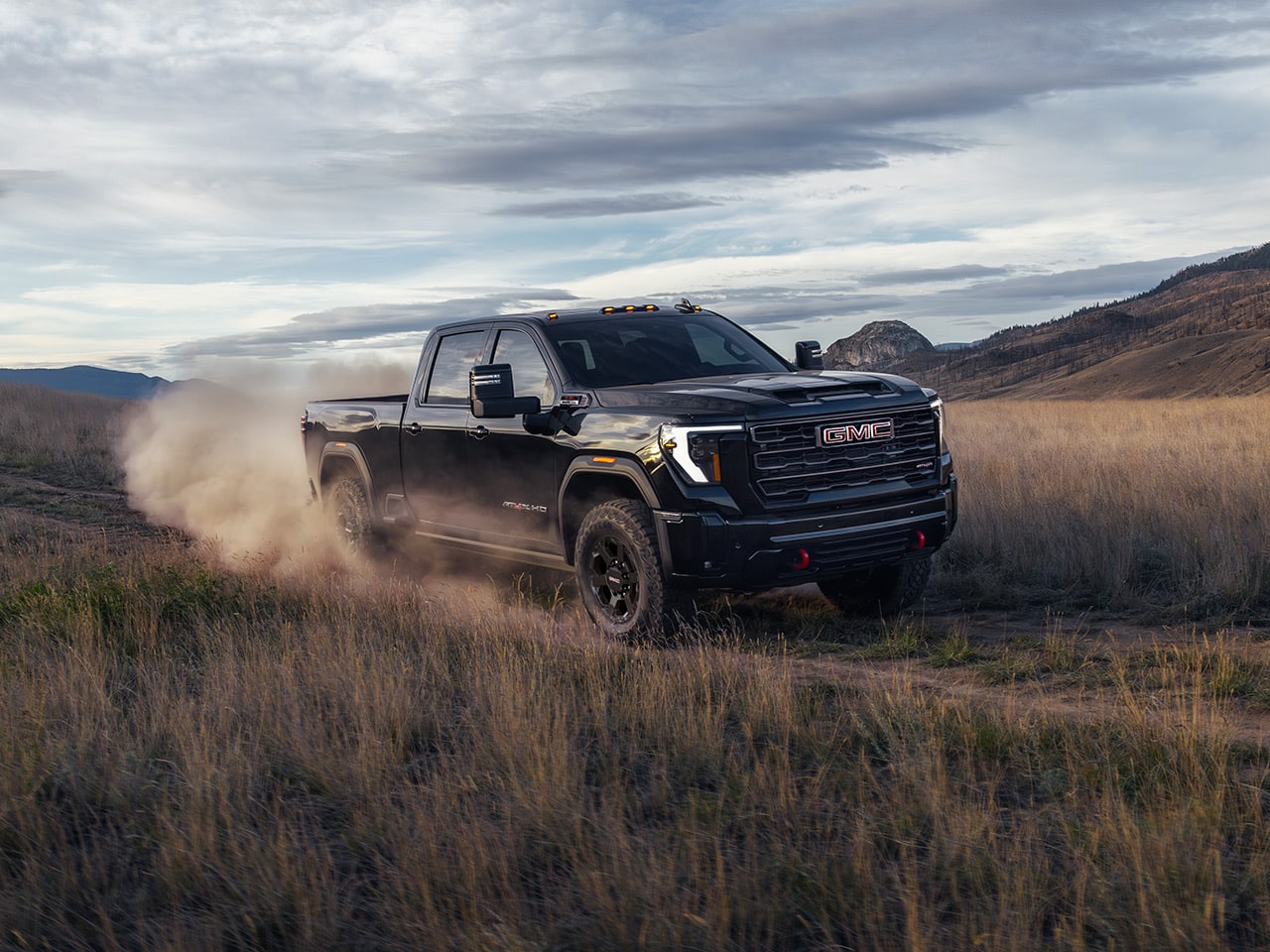 GMC Sierra HD 2027 noir roulant sur un chemin de terre et soulevant de la poussière dans une prairie sèche sous un ciel nuageux.