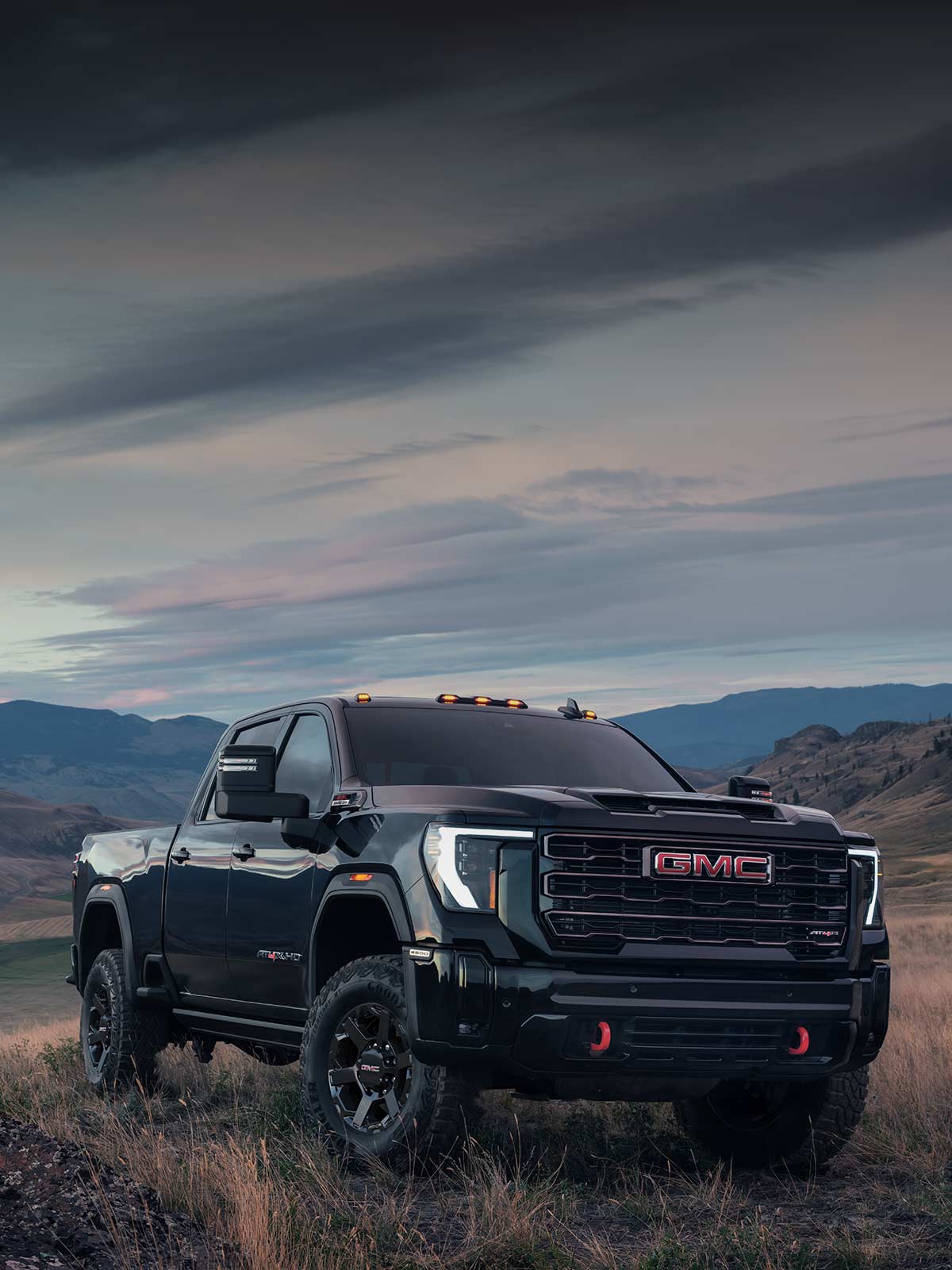 GMC Sierra HD AT4X 2027 gris foncé stationné sur une colline herbeuse, avec des montagnes sous un ciel nuageux.