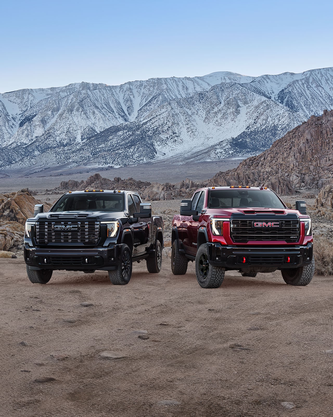 Two 2027 GMC Sierra HD Trucks, One Black and One Red, with Off-Road Tires and Red Recovery Hooks Parked Side By Side On a Rocky Terrain with Snowy Mountains in the Background 