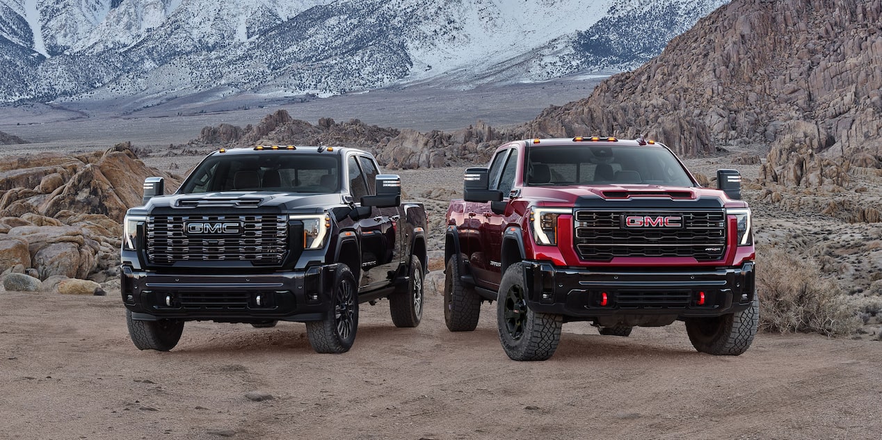 Two 2027 GMC Sierra HD Trucks, One Black and One Red, with Off-Road Tires and Red Recovery Hooks Parked Side By Side On a Rocky Terrain with Snowy Mountains in the Background 