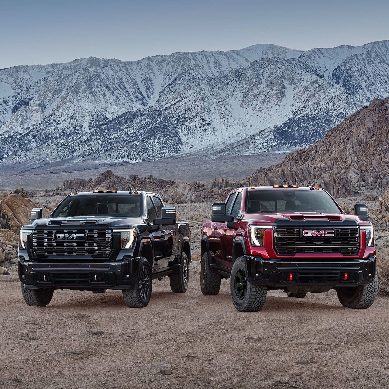 Two 2027 GMC Sierra HD Trucks, One Black and One Red, with Off-Road Tires and Red Recovery Hooks Parked Side By Side On a Rocky Terrain with Snowy Mountains in the Background