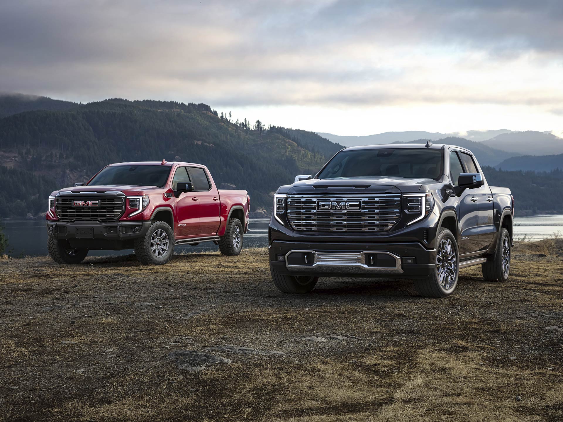 Two 2026 GMC Sierra 1500 Pickup Trucks Parked in a Grassy Field by a Body of Water.