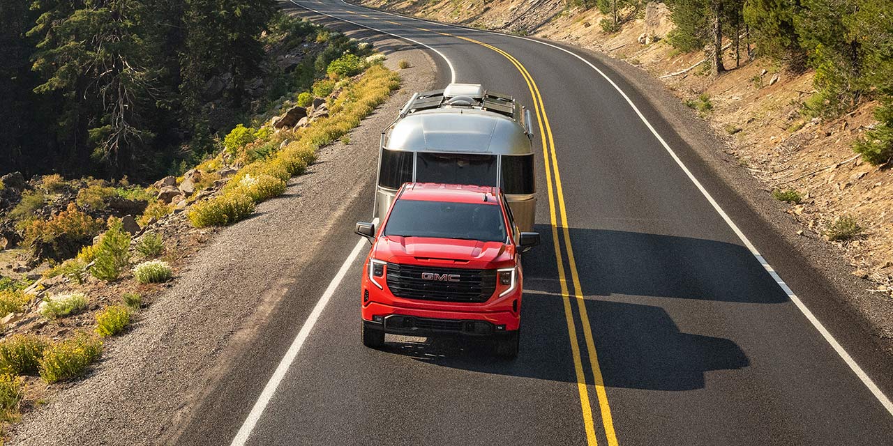 Birds-eye View of the 2026 GMC Sierra 1500 Towing a Trailer Down a Road Lined with Trees.