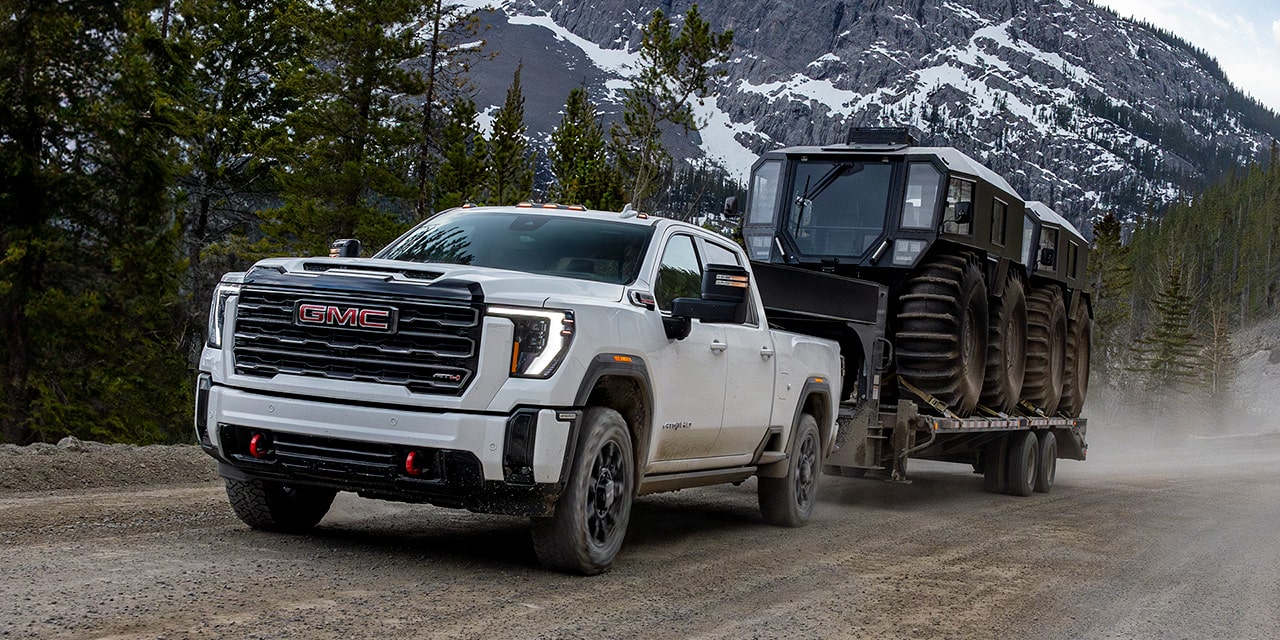 2026 GMC Sierra HD AT4 Pulling a Trailer Down a Dirt Road Lined with Pine Trees.