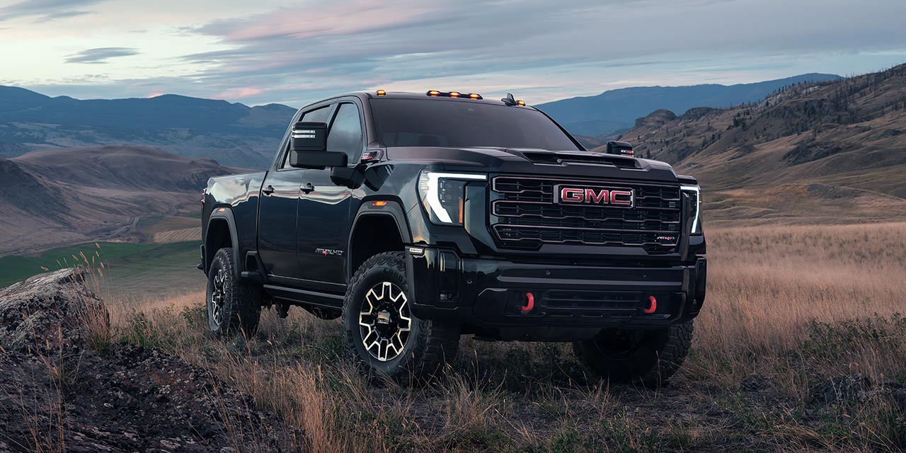 Front-Side View of a 2026 GMC Sierra HD Parked on a Terrain Field Surrounded by Mountains.